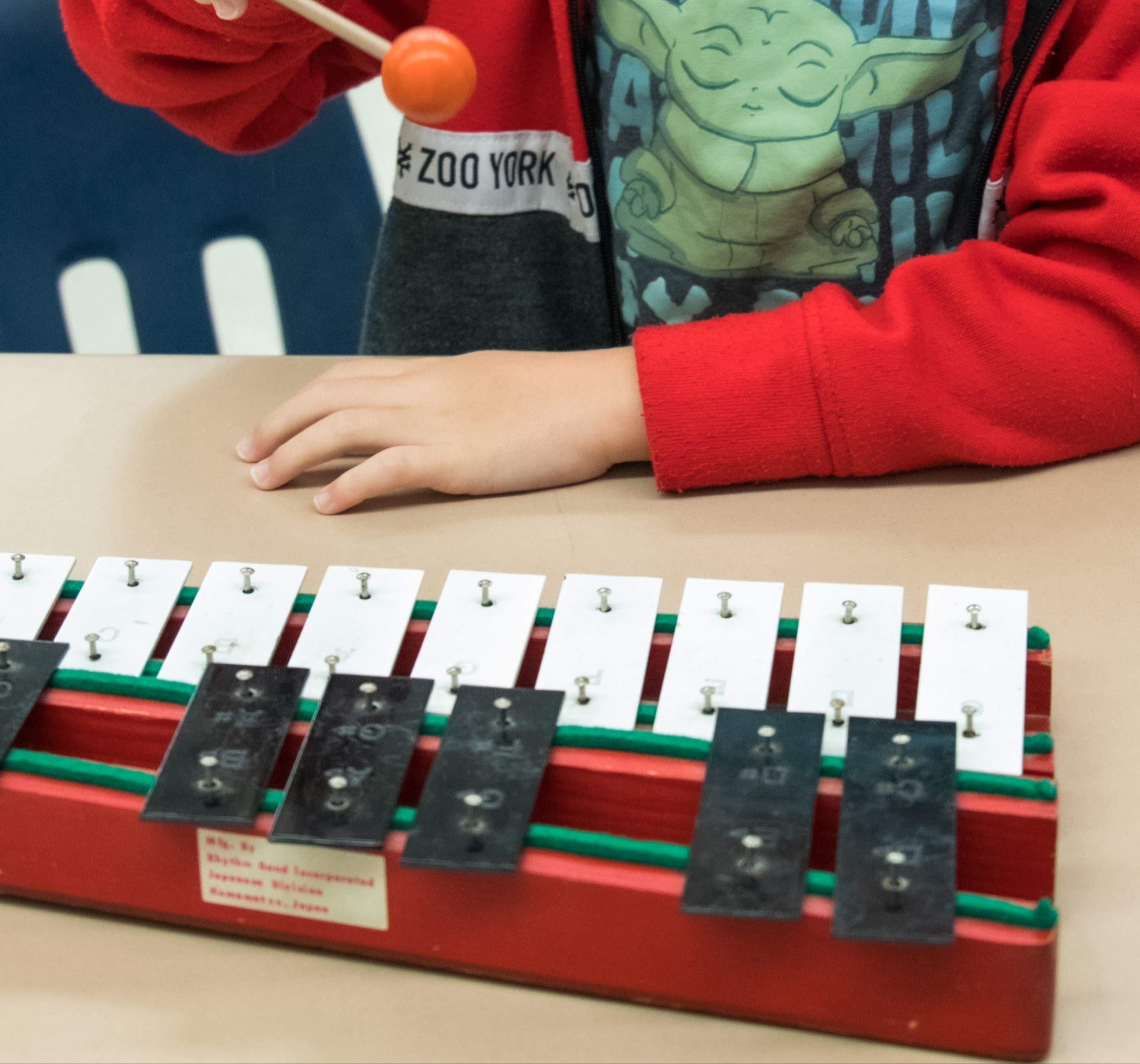 A child in a red jacket plays a xylophone with an orange mallet.
