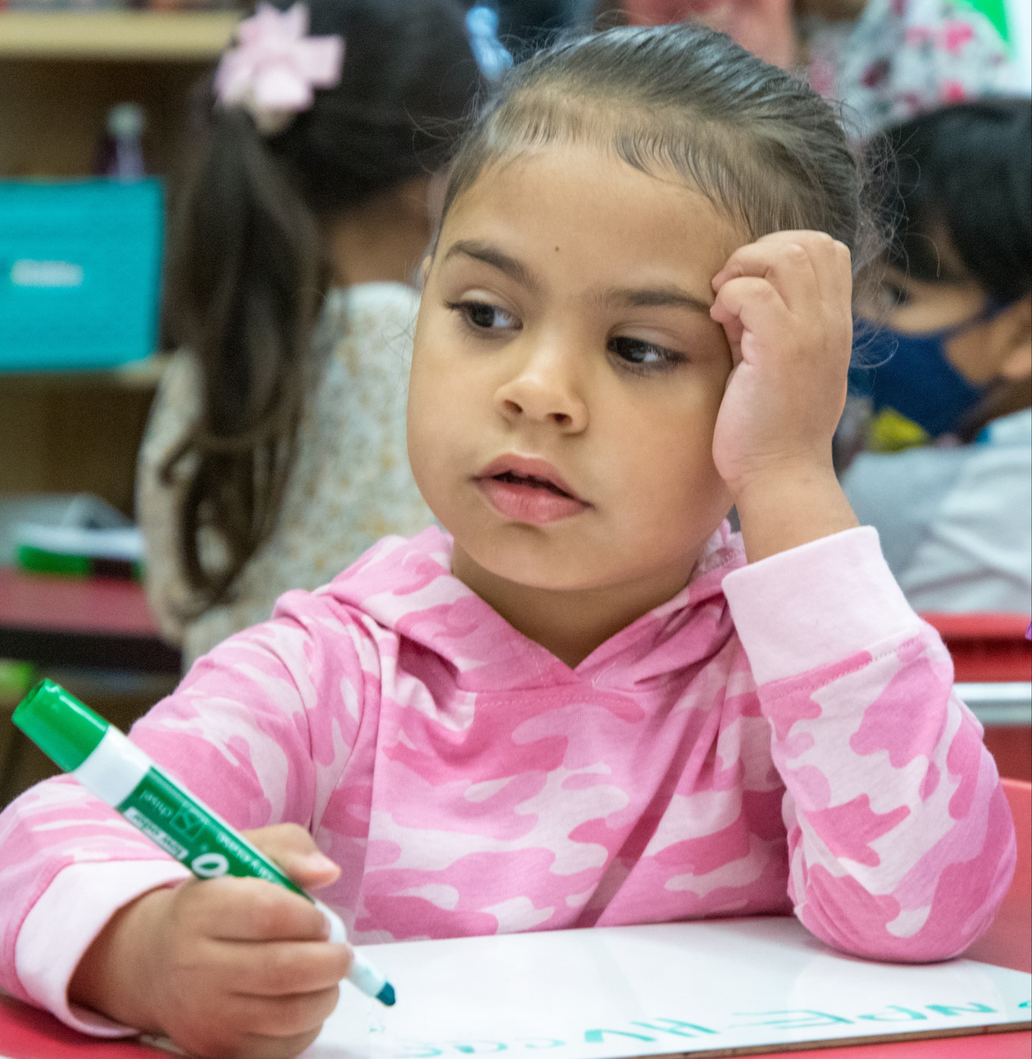A young girl in a pink hoodie sits at a desk, holding a green marker. She looks down at her paper.