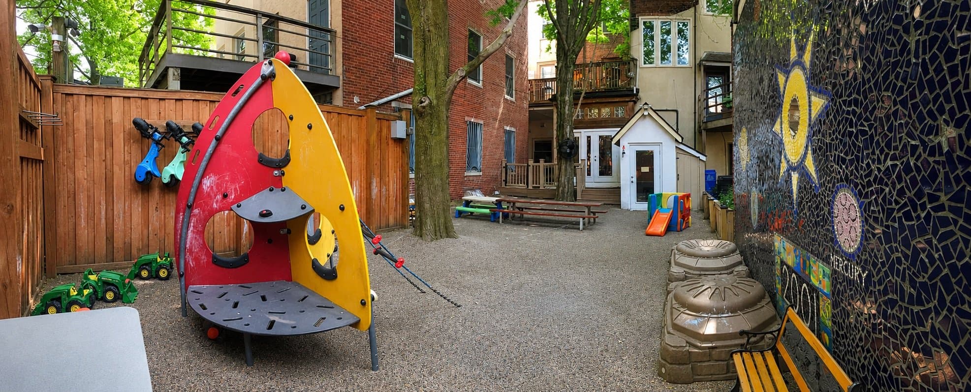 Enclosed outdoor preschool play area with climbing equipment, ride-on toys, picnic tables, and a colorful mosaic wall