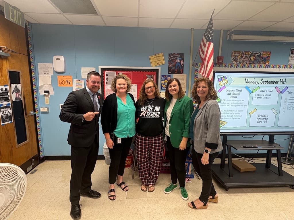 Five people, including a man in a suit, stand in a classroom. Two women wear green jackets, while two others wear black shirts.