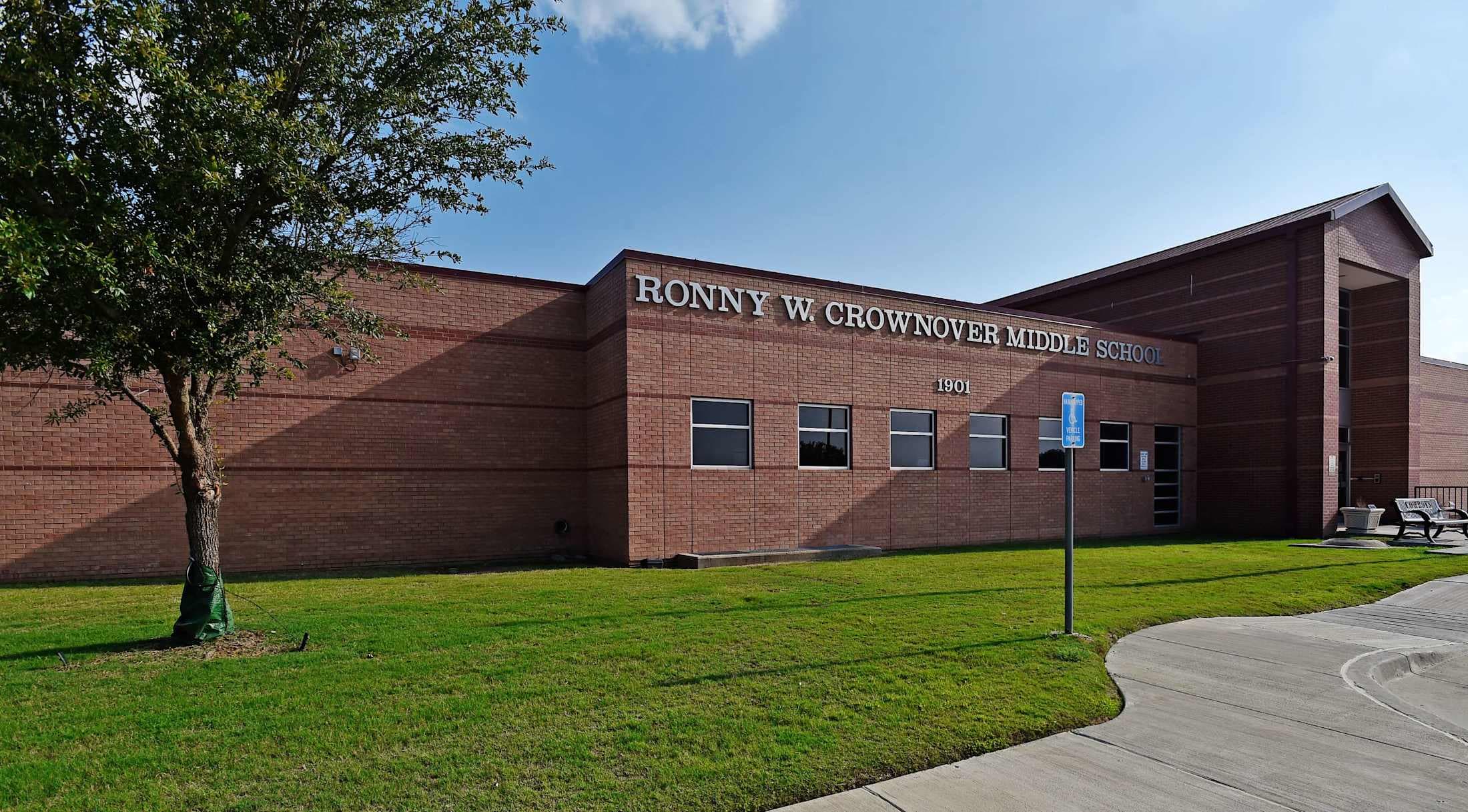 A brick building with the name %22Ronny W Crownover Middle School%22 displayed on the front, surrounded by a well-manicured lawn and trees in the foreground.
