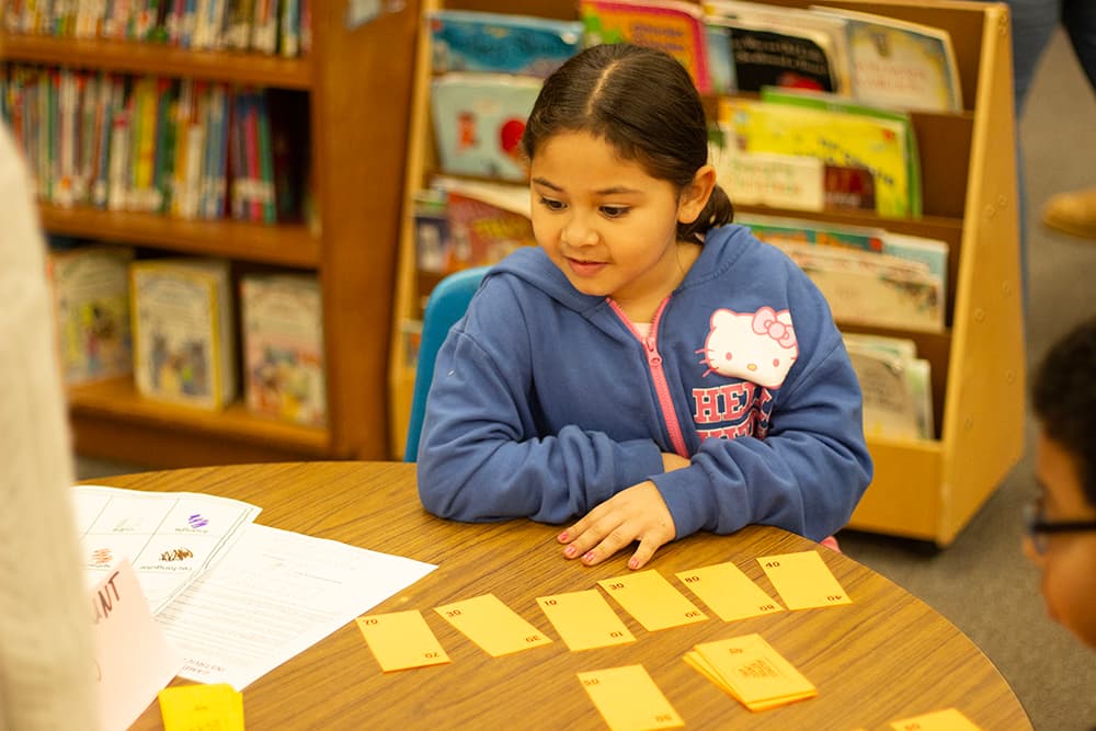 A young girl sits at a table with numbered flash cards in front of her as she works out a math problem.