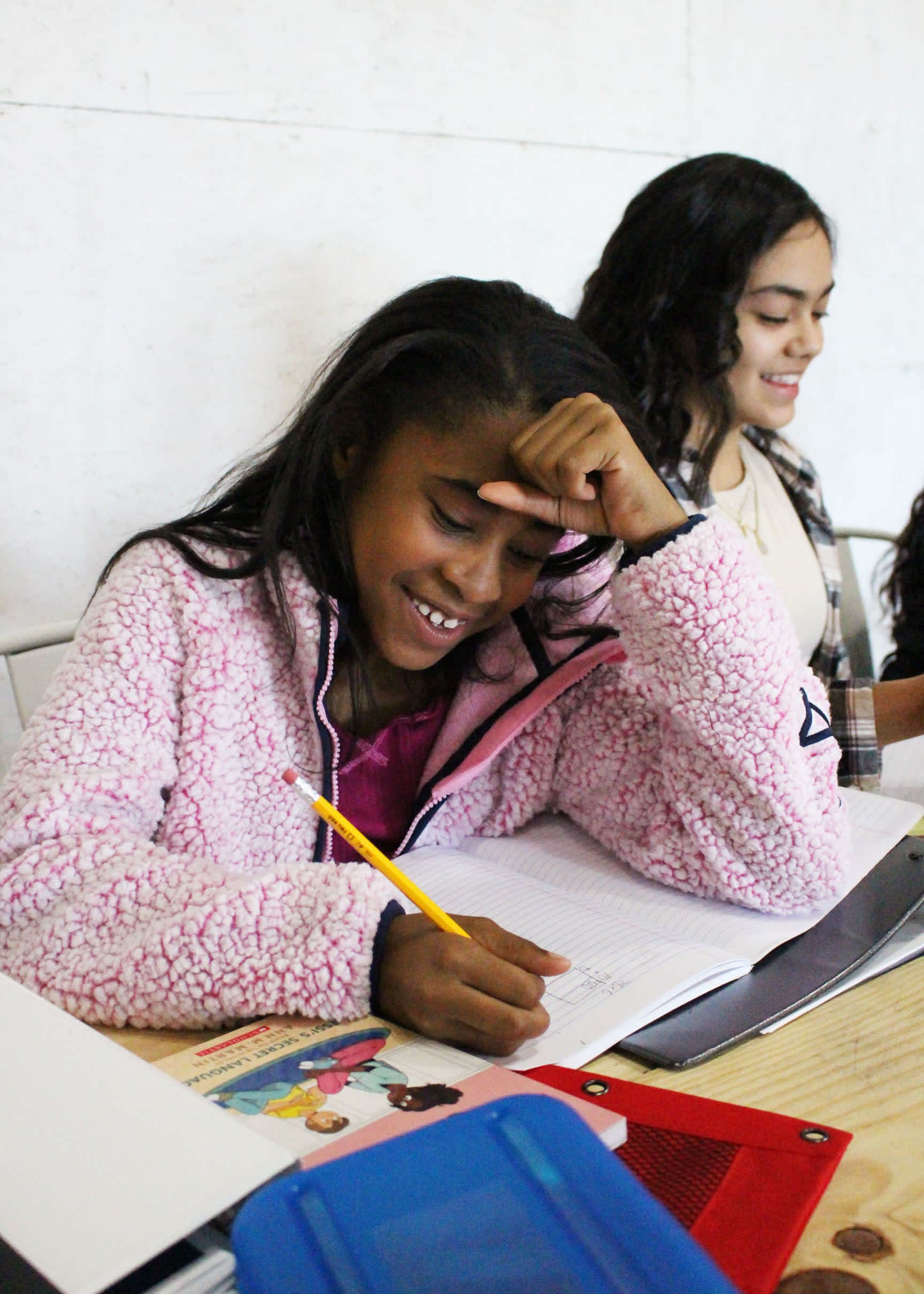 A young girl in a pink fuzzy jacket writes in a notebook at a desk, with another girl smiling in the blurred background.