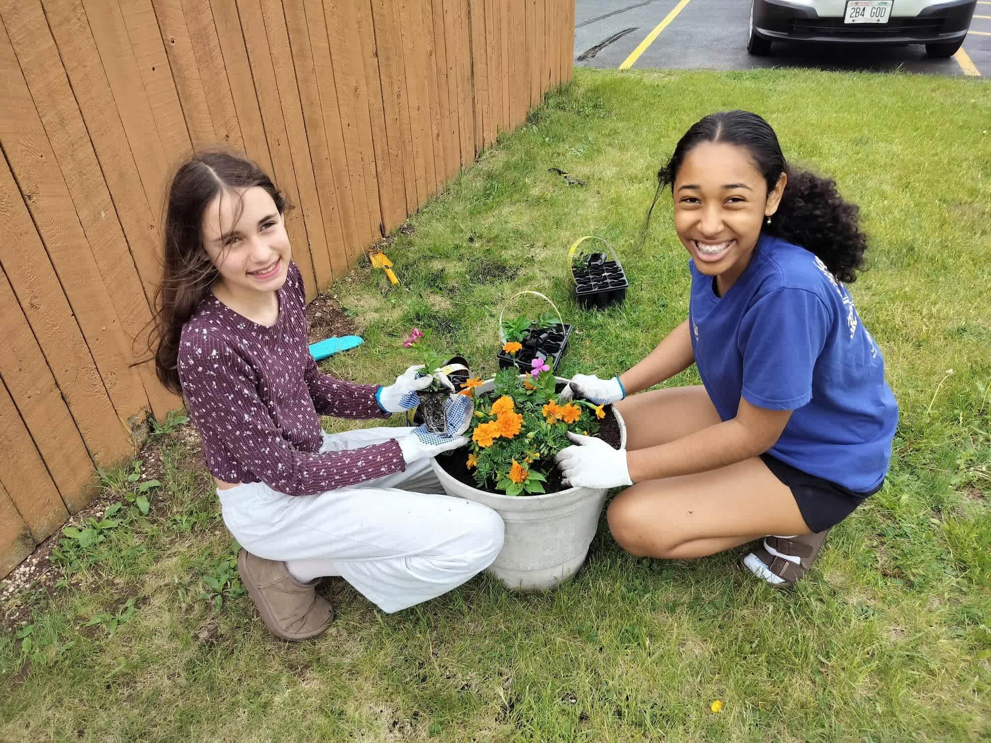 Two smiling girls in white gloves plant orange flowers in a pot on a grassy lawn, with a wooden fence and a parked car in the background.