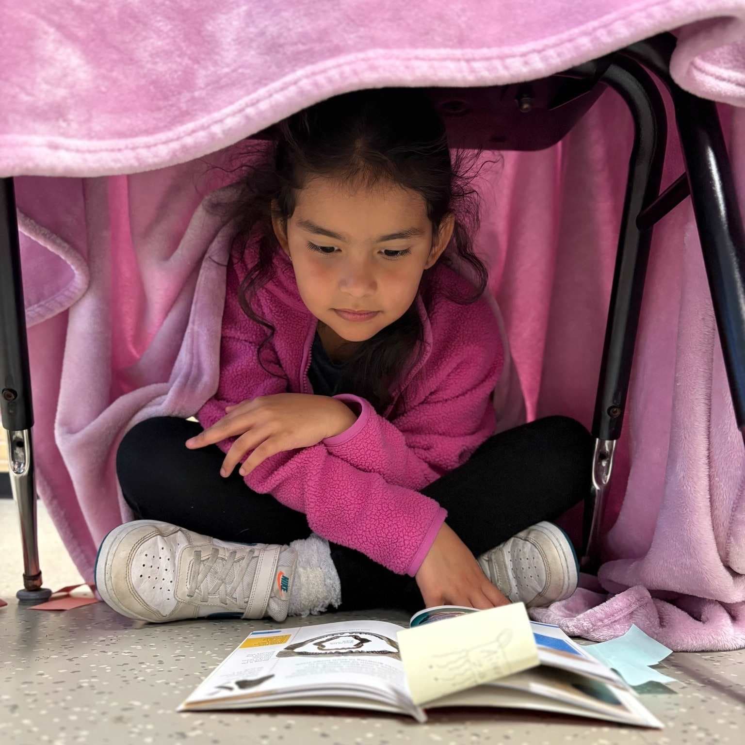 A young girl sits cross-legged on the floor, engrossed in reading a book, surrounded by a cozy pink blanket and a comfortable-looking environment.