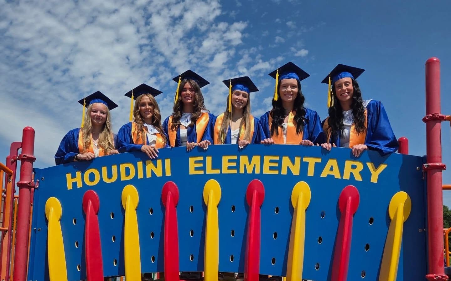 Five graduates in caps and gowns stand atop a colorful playground structure with %22Houdini Elementary%22 written across it, against a bright blue sky with scattered clouds.
