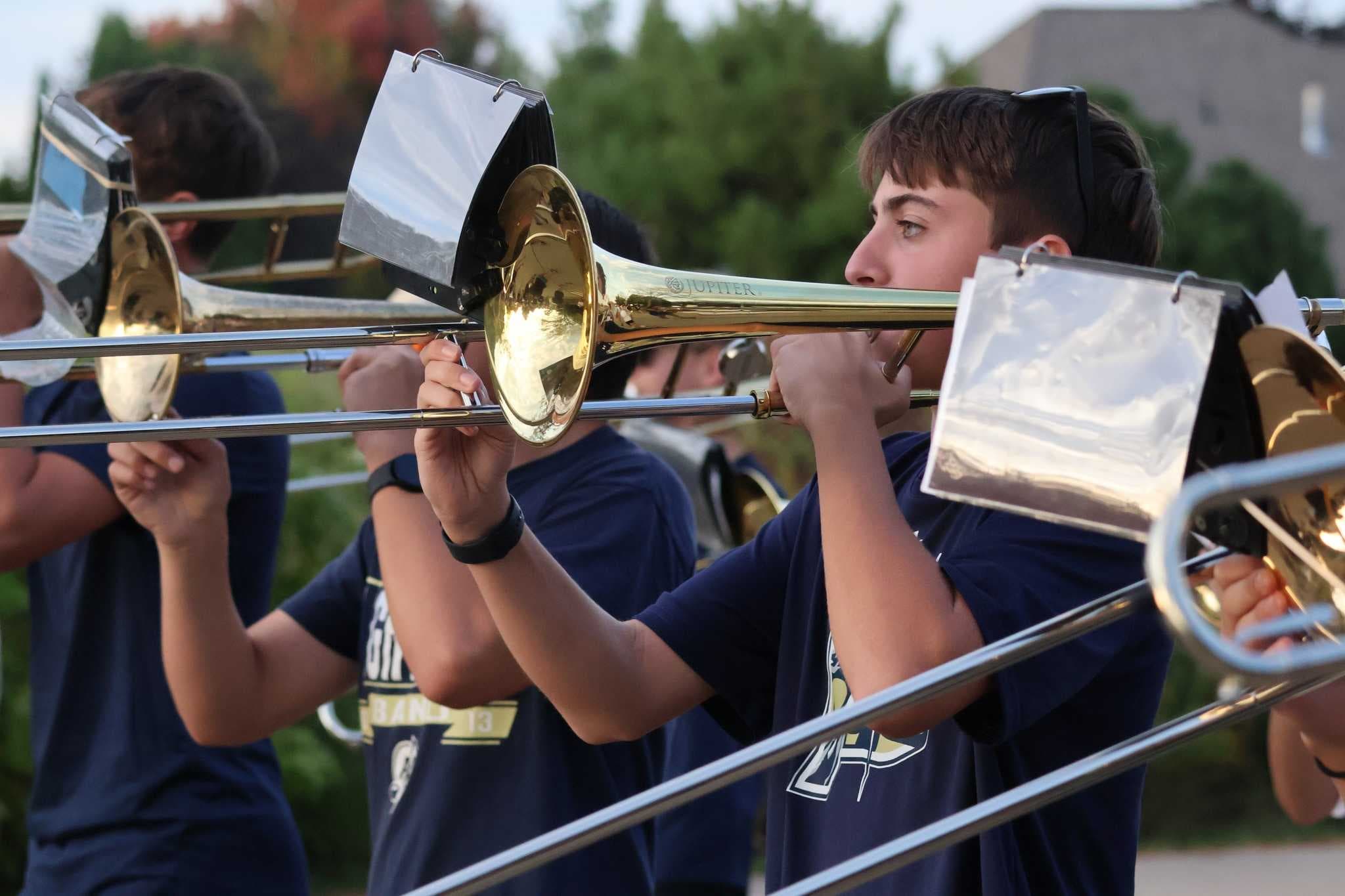 A group of students in school uniforms playing brass instruments outdoors, with a forested background.