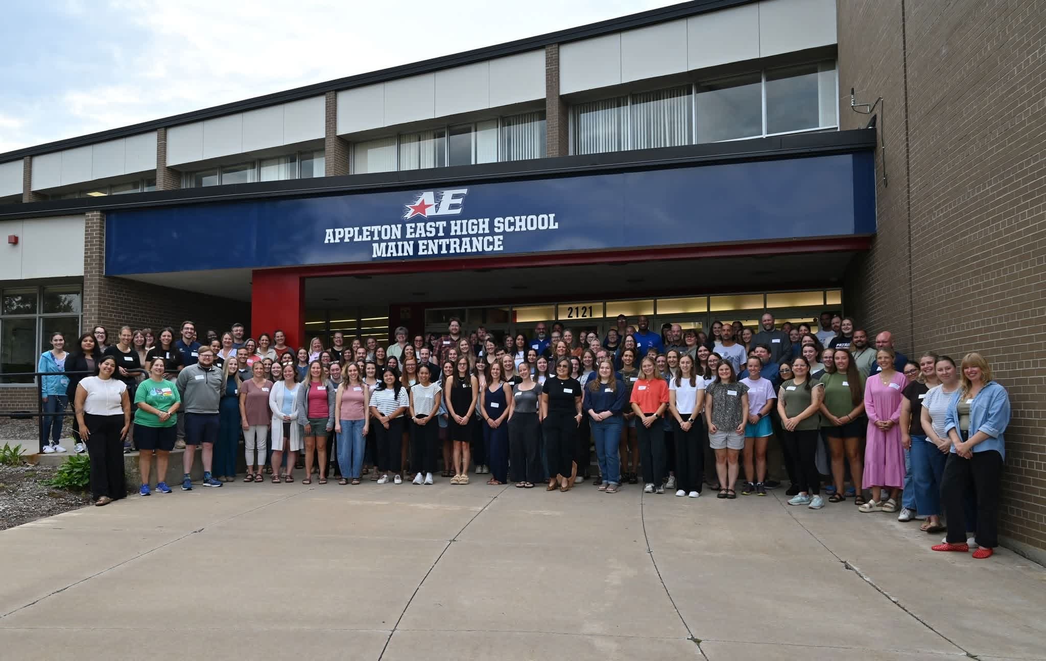 A large group of people stands on a paved walkway in front of the brick building of Appleton East High School.