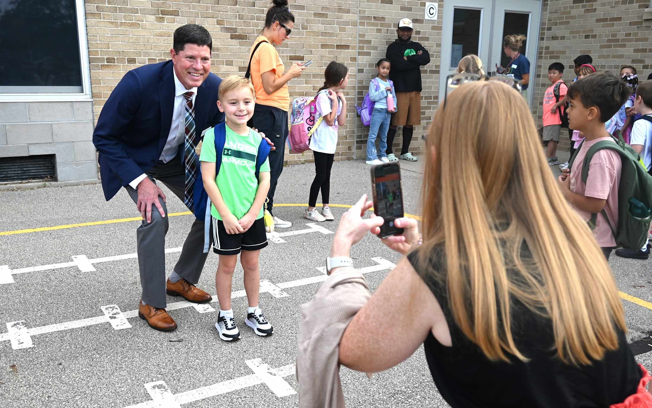 A woman photographs a young boy in a green shirt and a man in a suit posing on a marked asphalt area, with other children and adults in the background near a brick building.