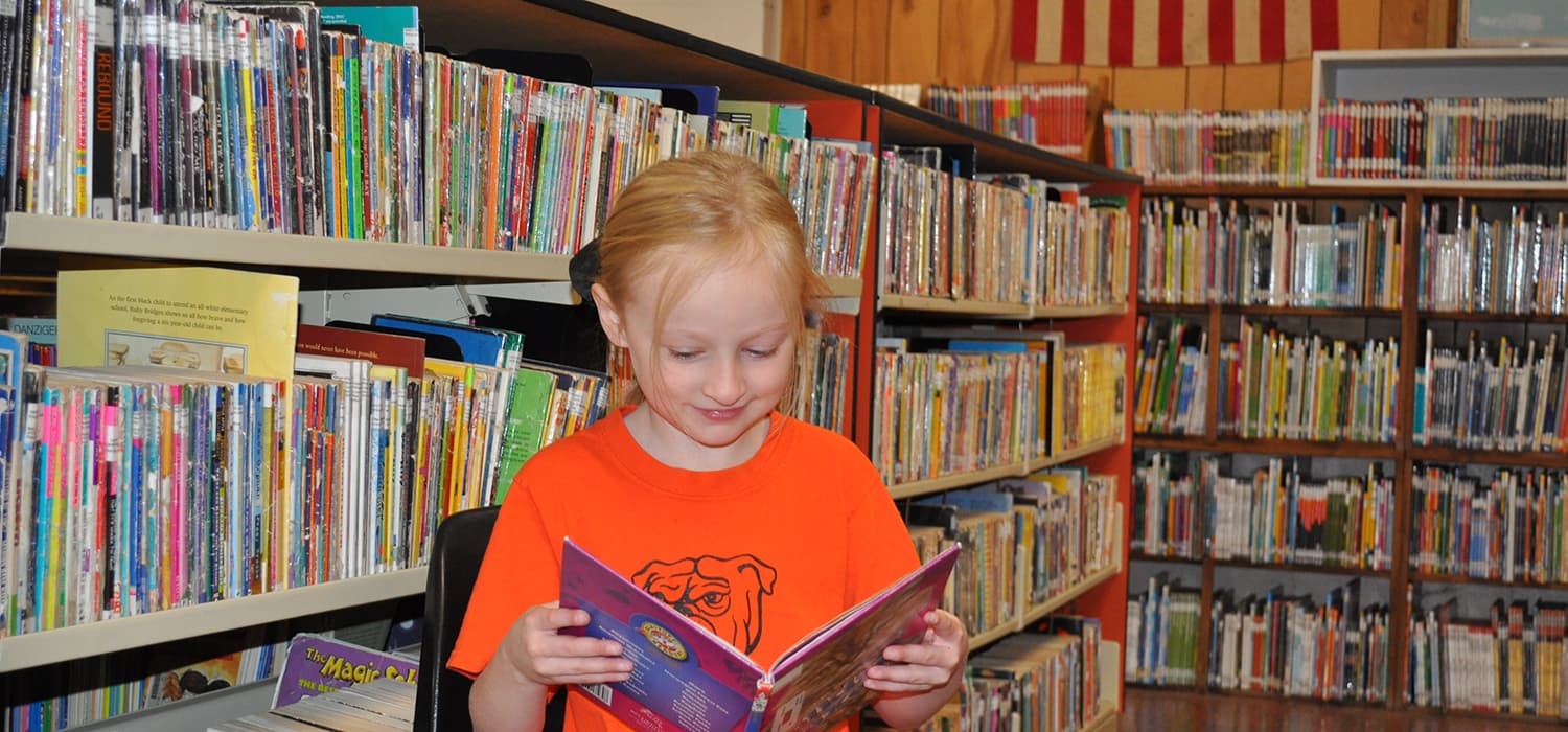 A young child smiles while holding a book in a library.