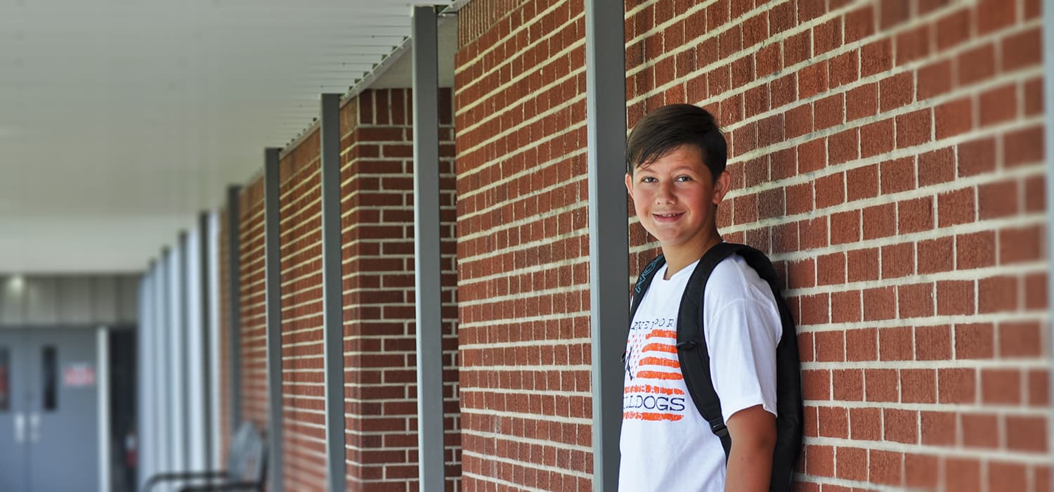 A young person with a backpack stands near a brick wall, looking at the camera.