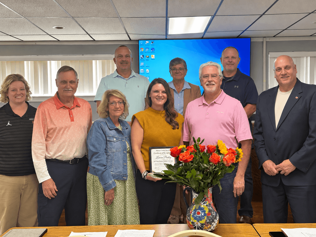 A group of people stands around a table with a flower arrangement and certificate. A woman in the center holds the certificate.