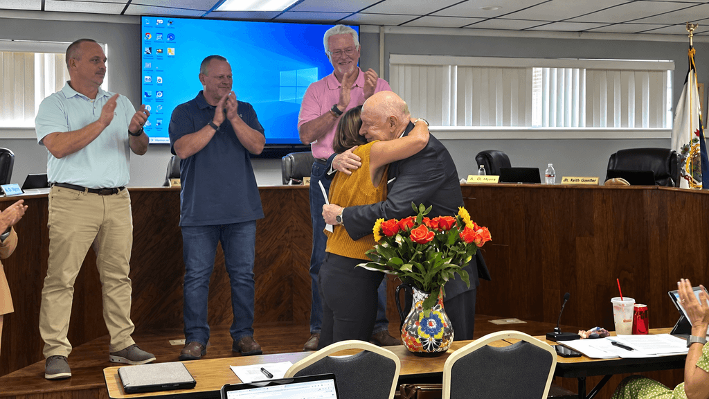 Group of people in a room. A man hugs a woman. A podium is in the foreground.