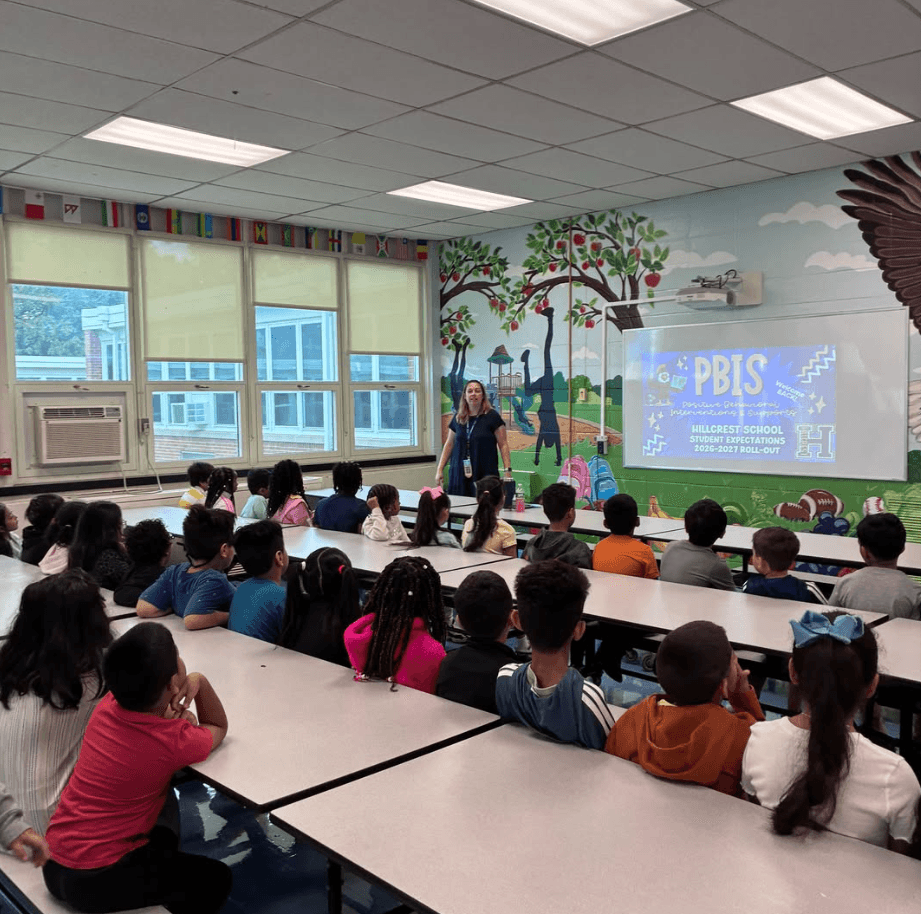 Classroom with painted walls; a teacher stands in front of a projector screen. Students seated at tables listen.