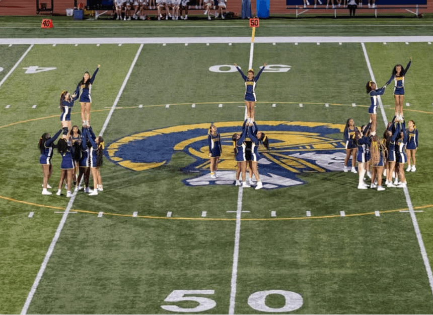 Group of cheerleaders on a football field performing stunts; school logo at center.