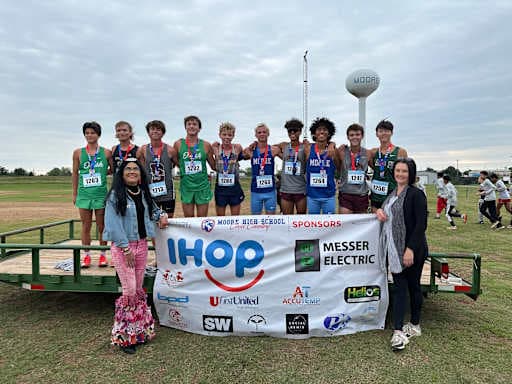 A group of young women in athletic wear and medals stand behind a banner with sponsors' logos, while two adults stand in front of them, with a water tower visible in the cloudy background.