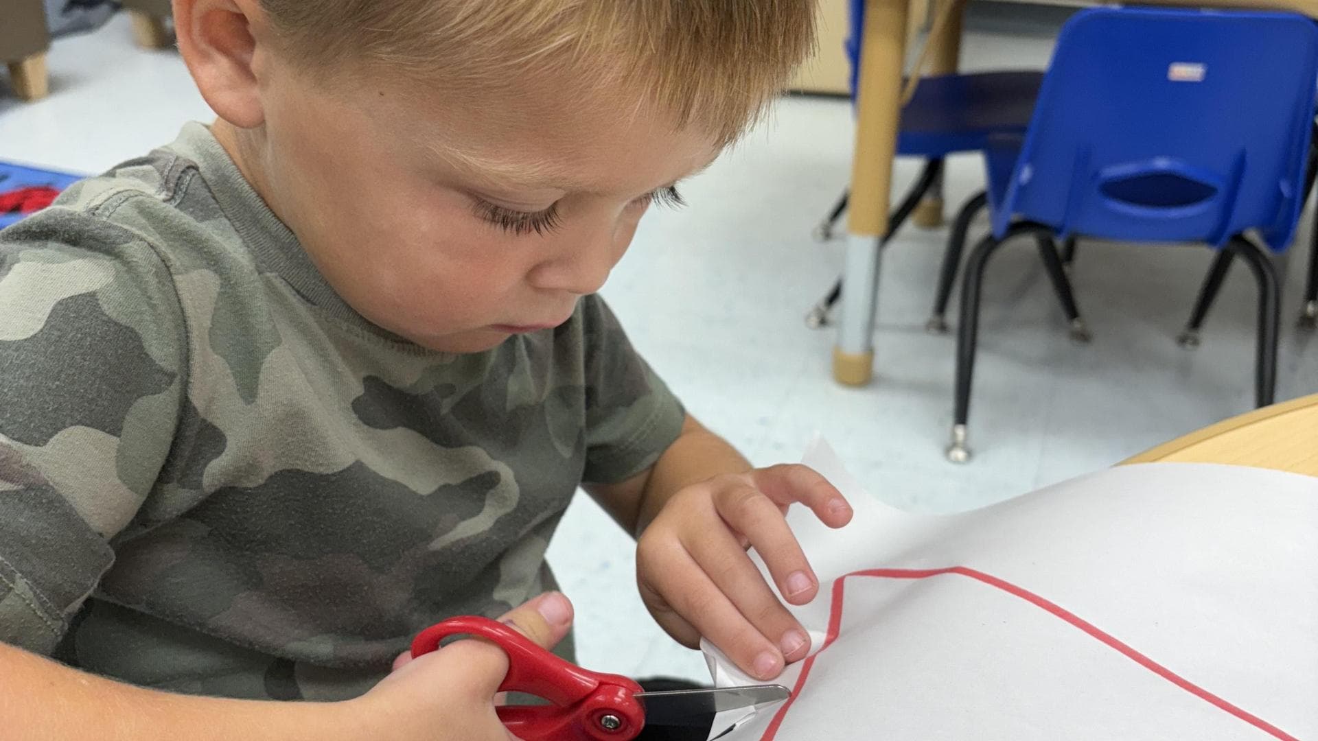 Young child using scissors to cut a red shape from white paper.