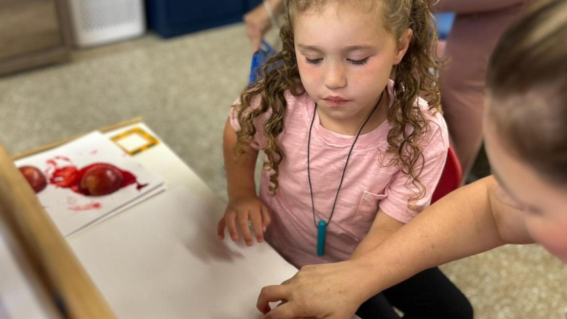 A young girl with curly hair focused on art, with an adult's hand assisting her.
