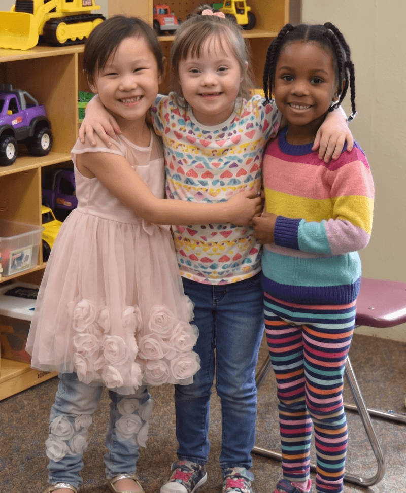 Three young girls, two smiling widely and one with Down syndrome, embrace each other warmly.