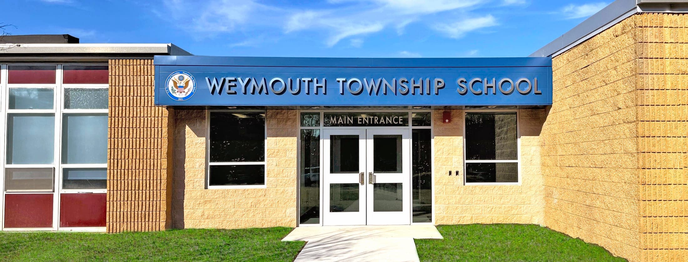 A bright blue sign above the main entrance of Weymouth Township School is set against a tan brick building, with a clear blue sky and white clouds in the background.