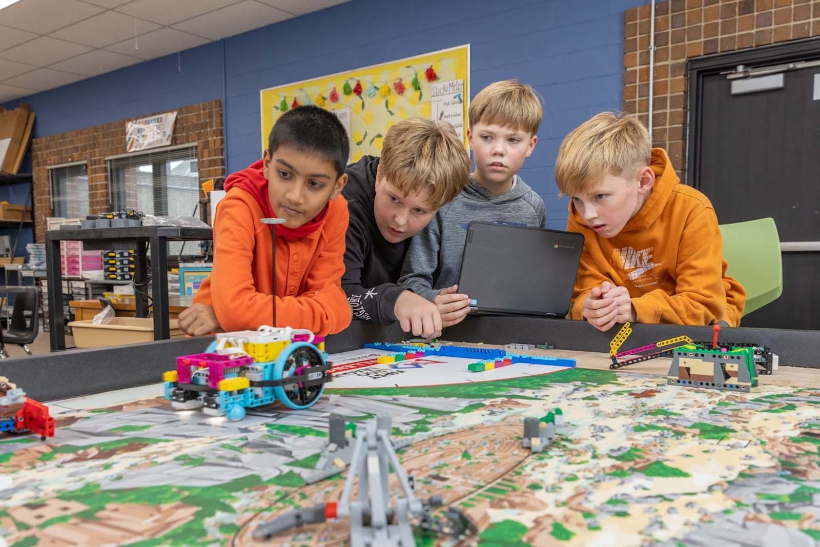 A group of young students, dressed in colorful clothing, are gathered around a table examining a model or toy, with a vibrant, art-filled classroom environment visible in the background.