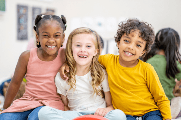Three diverse children smile and embrace, looking happy.