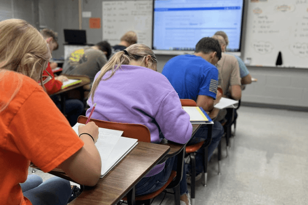 Students are seated at desks in a classroom, working on assignments.