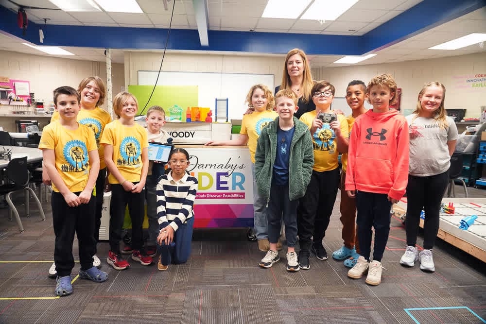 A group of smiling children and an adult stand in front of a colorful display in a classroom.