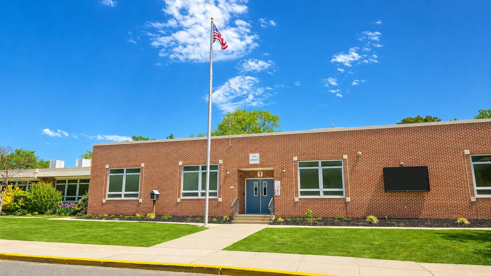 A brick school building with a flagpole in the foreground stands against a bright blue sky with fluffy clouds, surrounded by green grass and trees.