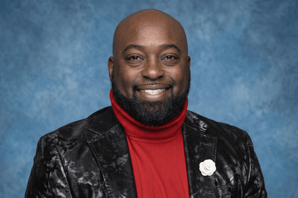 A smiling Black man with a beard wears a red turtleneck and a patterned black jacket.