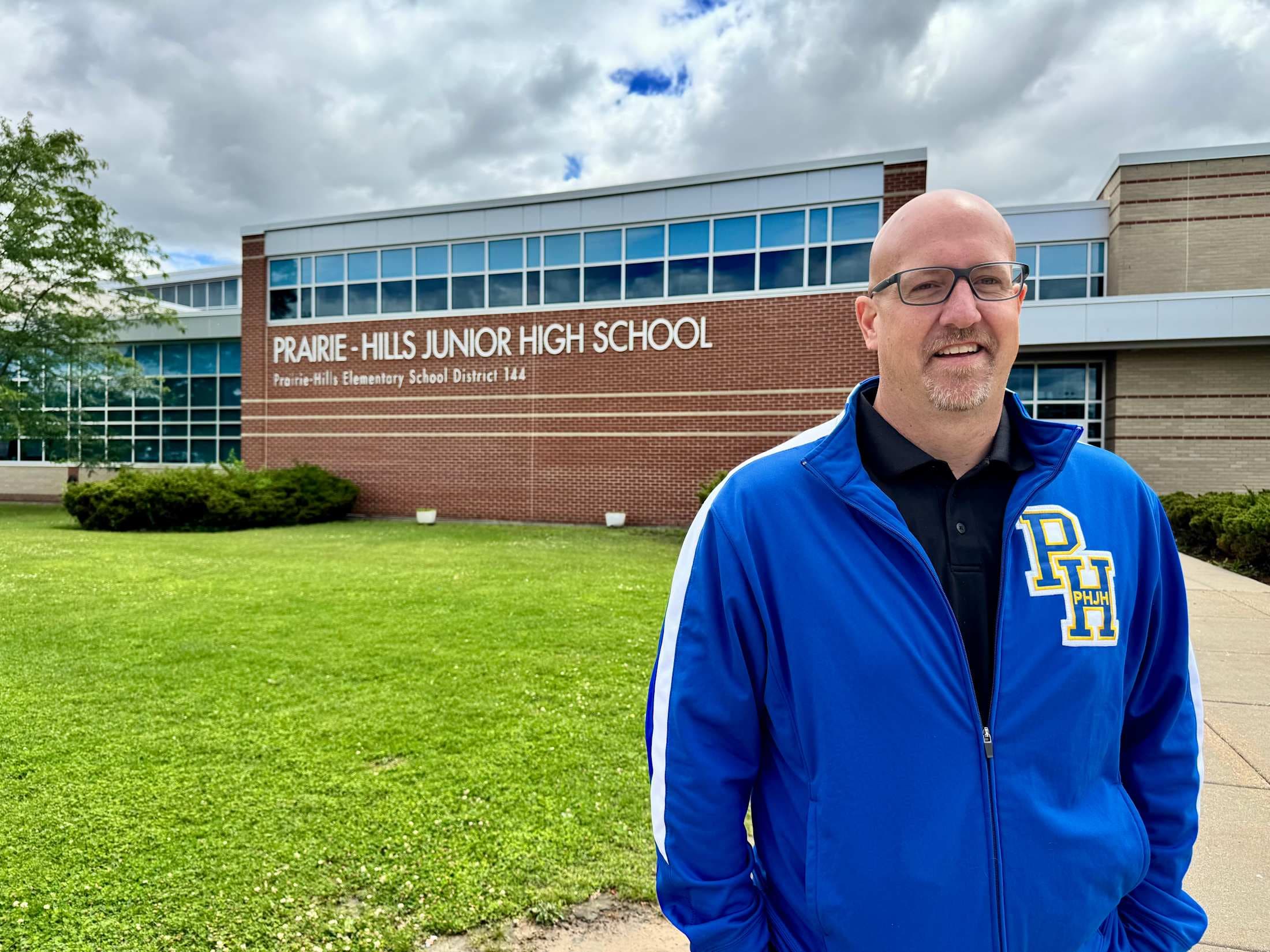 A man in a blue jacket stands in front of Prairie Hills Junior High School with a grassy lawn in the foreground and a cloudy sky in the background.