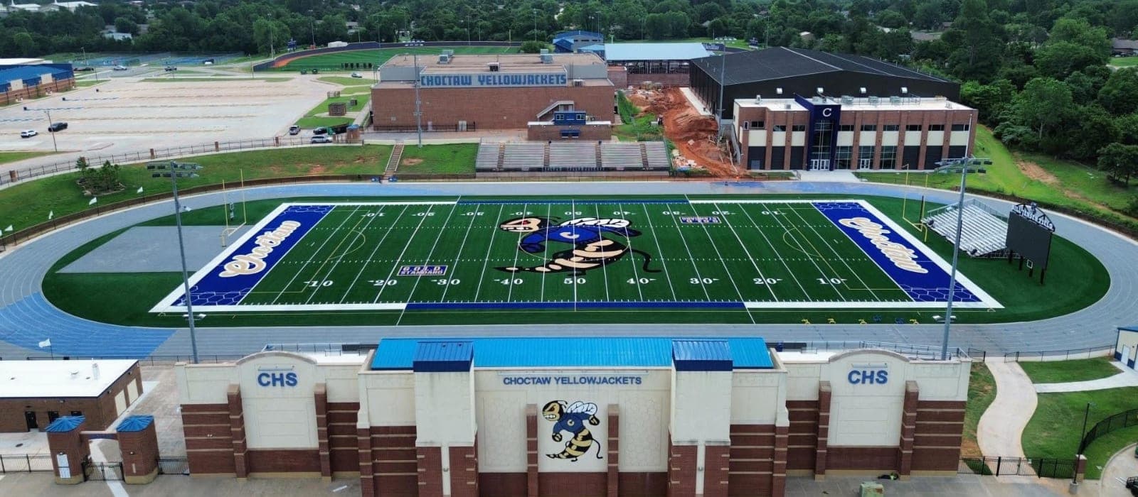 A football stadium with a blue and green field and track is in the foreground, while a school building and trees are in the background.