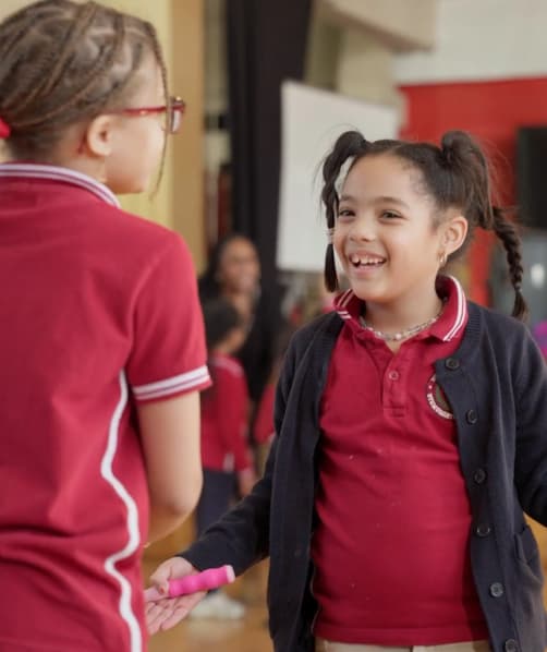 Two young girls in maroon school uniforms holding hands and smiling indoors.