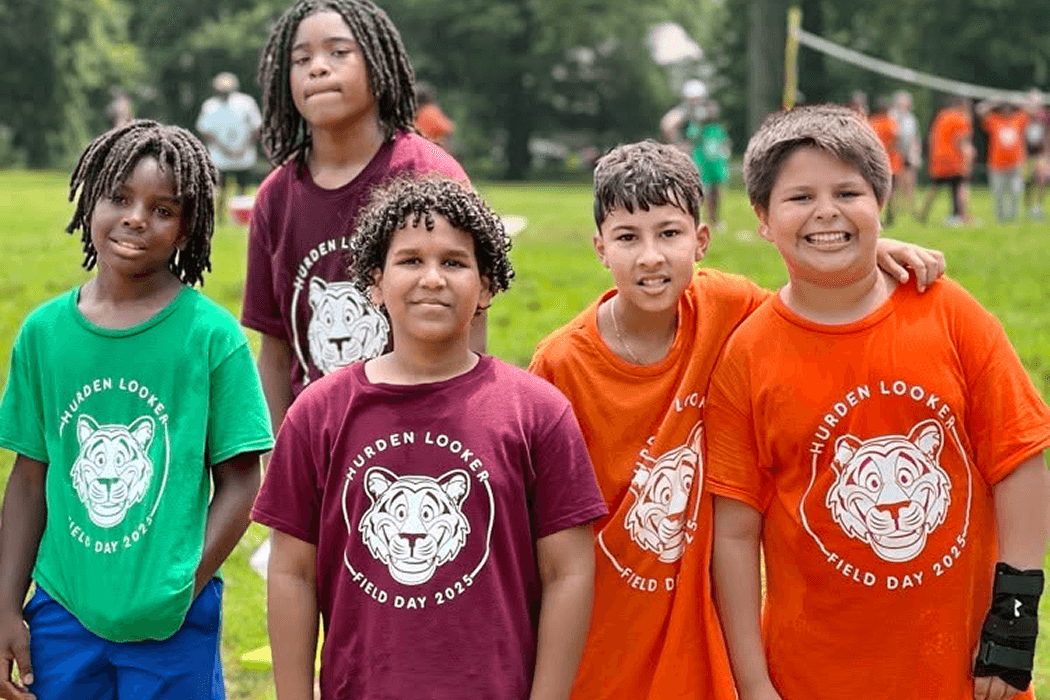 Five smiling children in colorful t-shirts stand together outdoors.