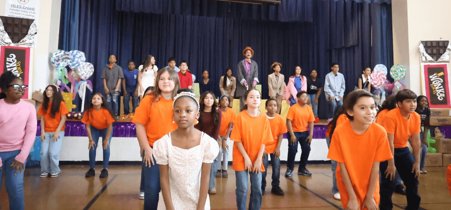 A group of children in orange shirts stand on a stage in front of an audience.