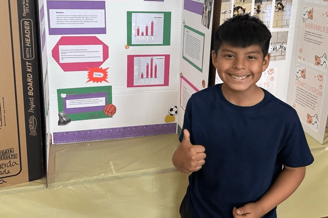 A young boy smiles and gives a thumbs-up next to a colorful science fair project board.