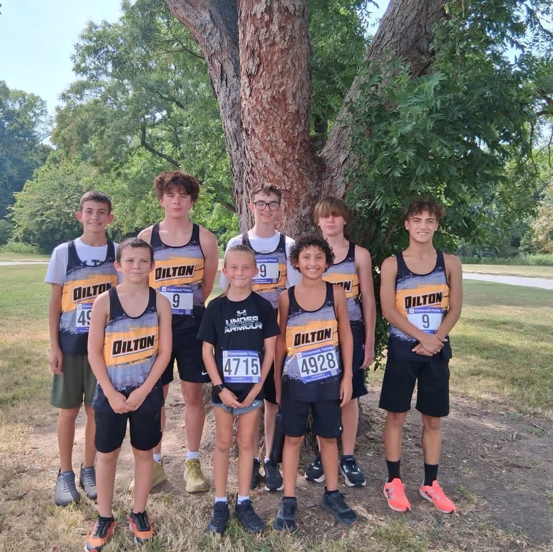 A group of eight young runners in matching uniforms pose together in front of a large tree outdoors.