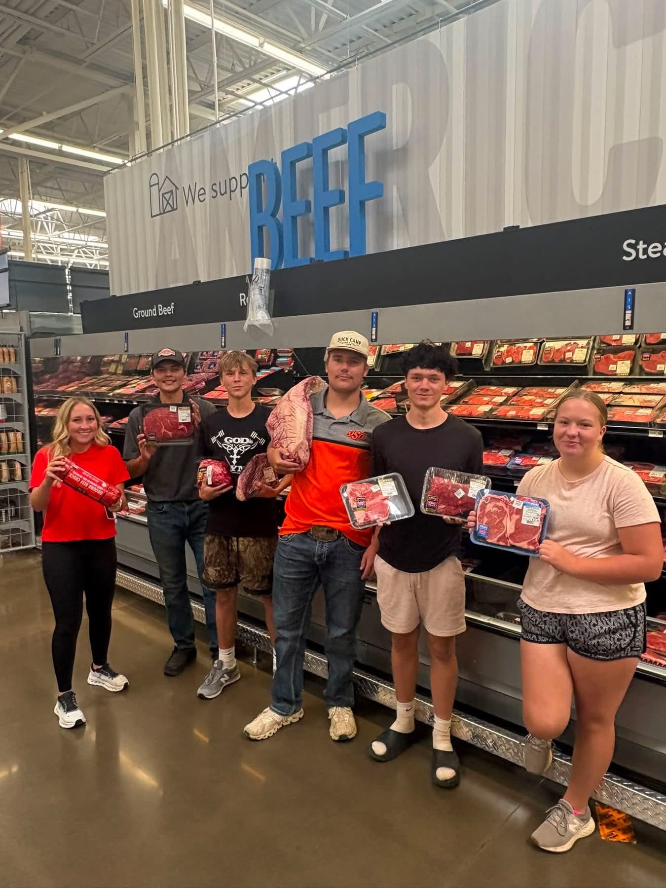 A group of five smiling young people pose in front of a supermarket's beef display, holding packages of meat.