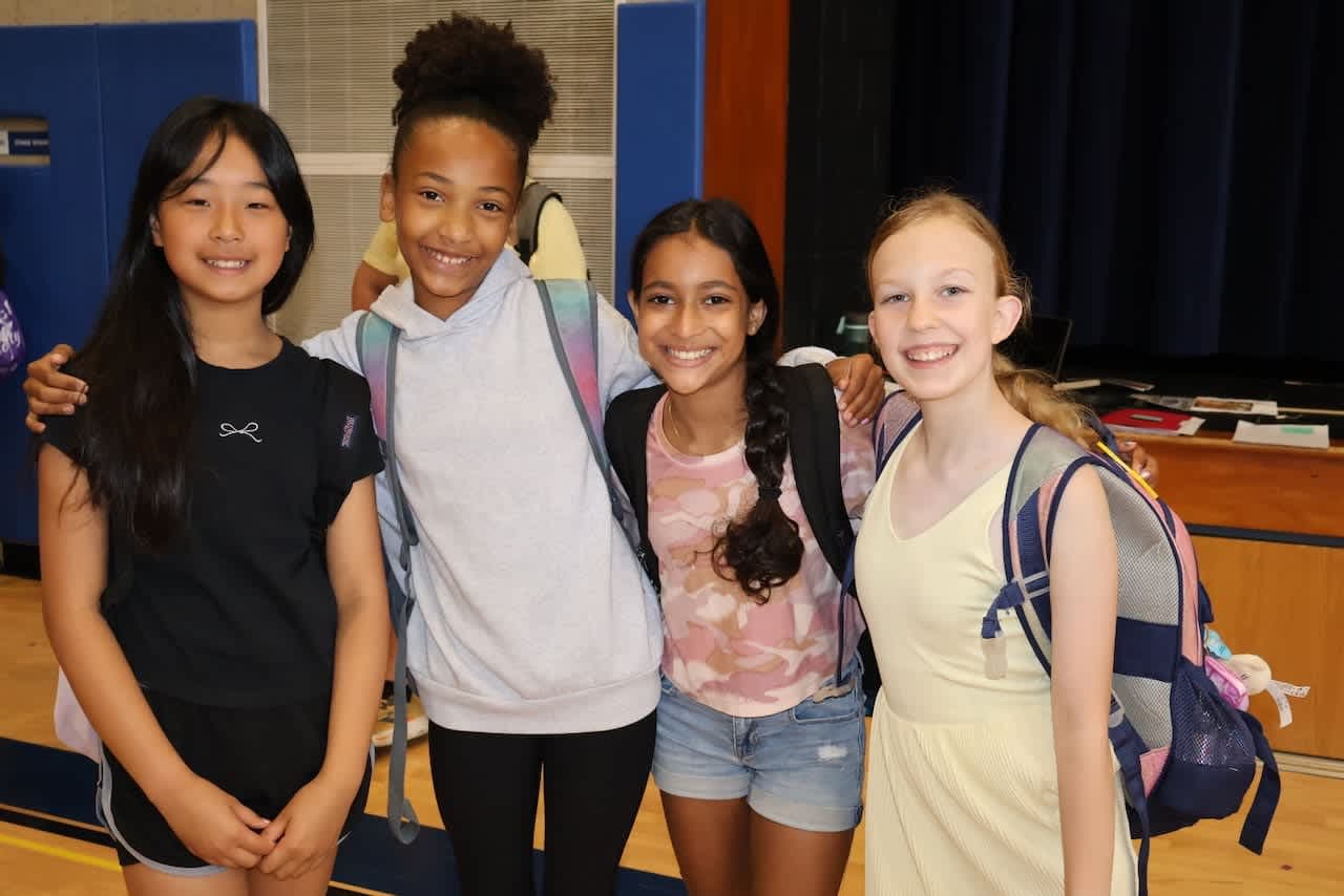 Four girls wearing backpacks, smile and pose for a photo in a school gym.