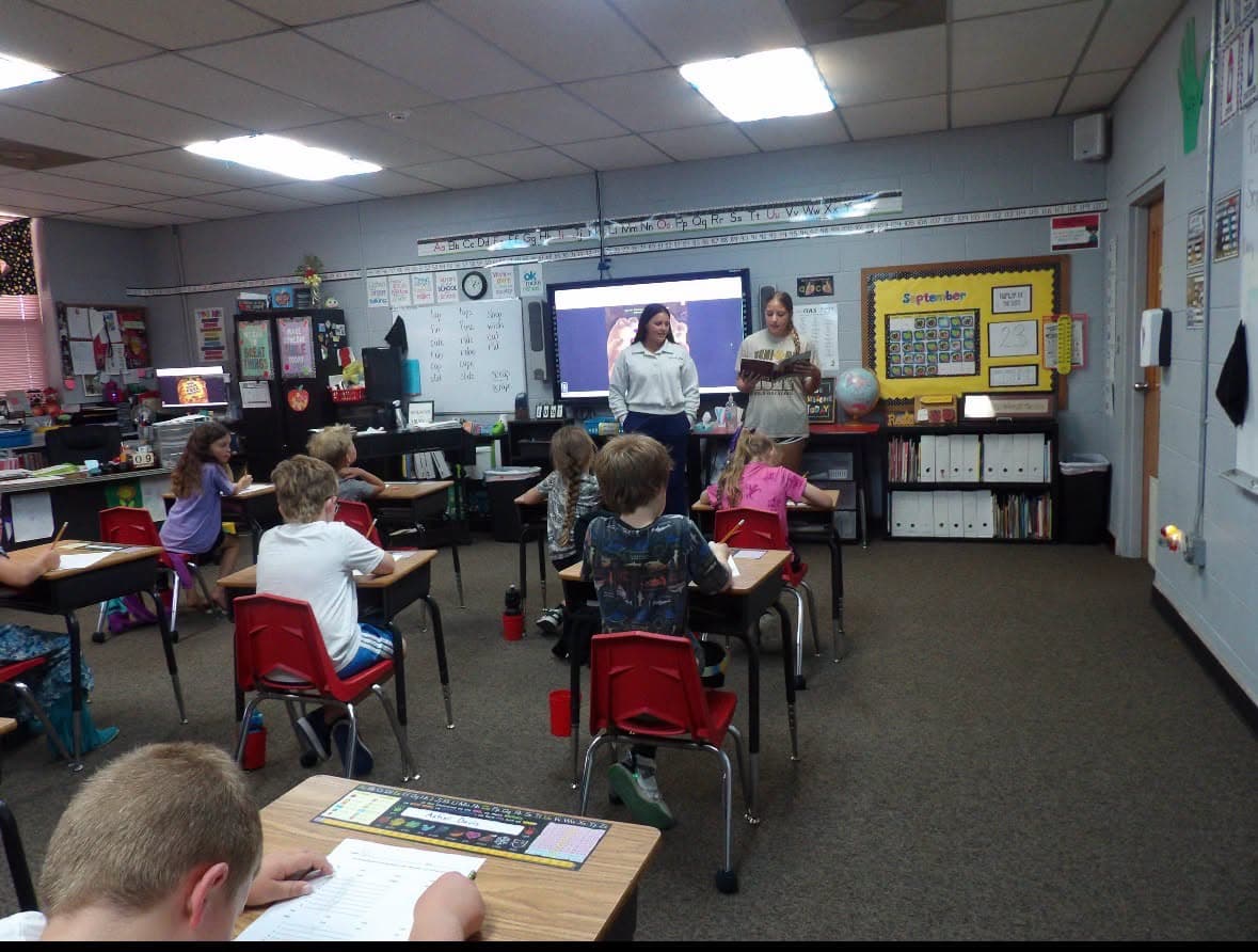 Students sit at desks in a brightly lit classroom, facing a digital screen.