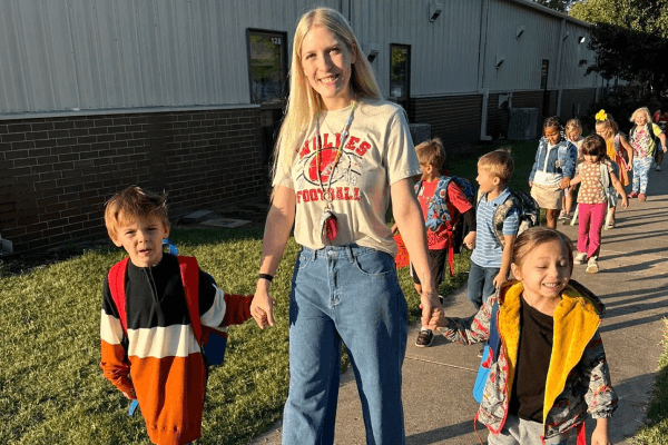 A teacher leads a group of young students in a line, holding hands as they walk outside a school building.