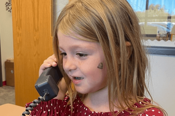 A girl in a red shirt with white polka dots holds a phone to her ear.