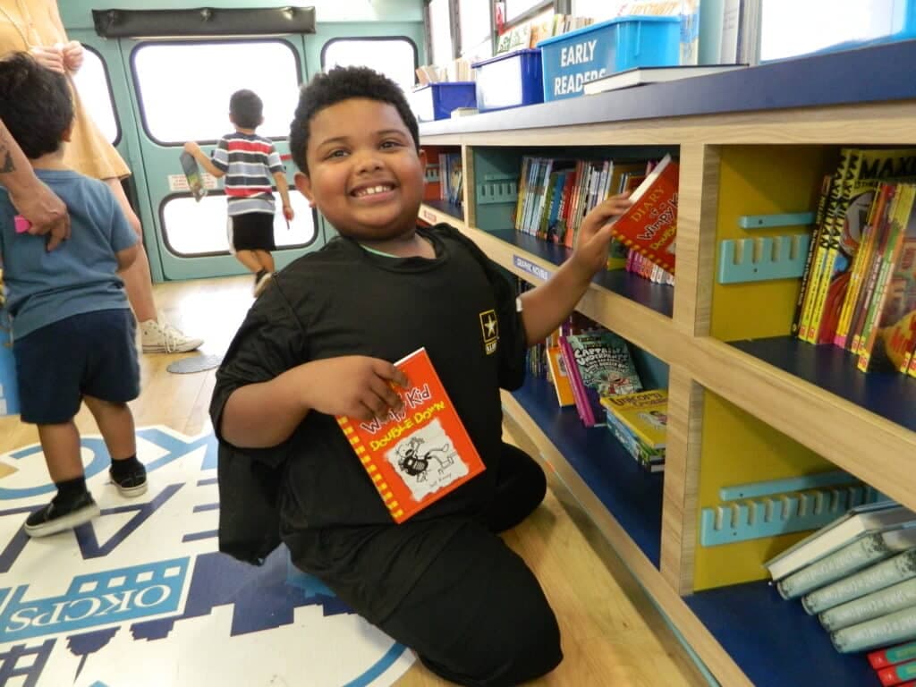 A smiling boy in the foreground holds a book while reaching for another on a shelf in a brightly lit library with other children in the background.