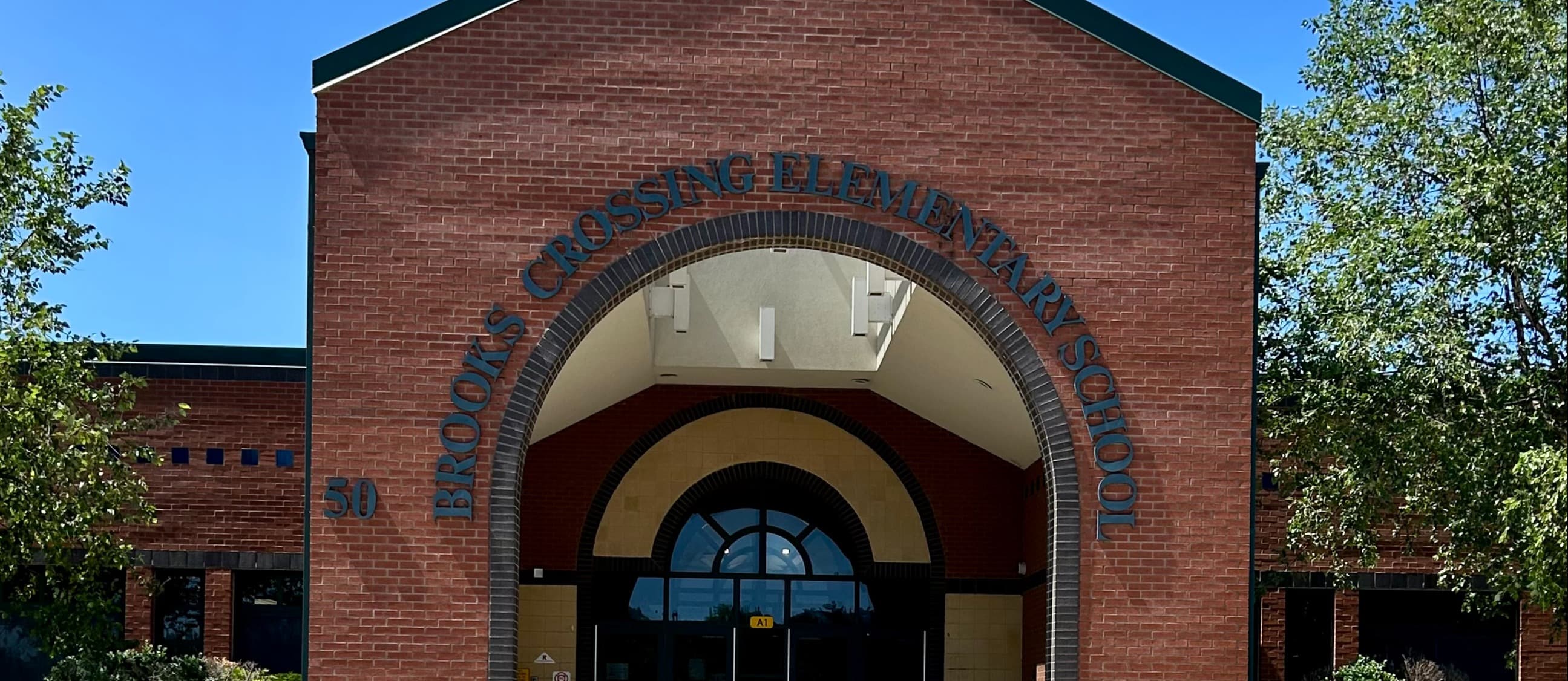 A brick building entrance with an arched doorway. The archway has glass windows and a door. Trees and shrubs surround the entrance.