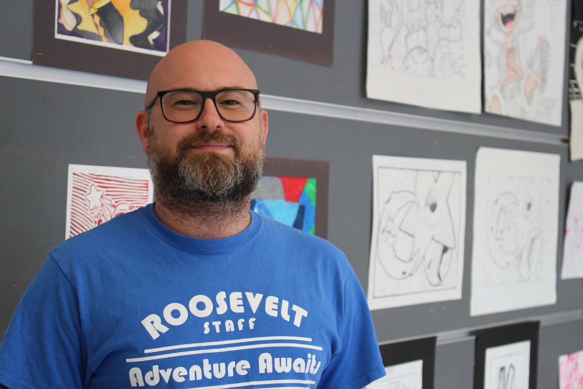 A bald man with glasses and a beard smiles in the foreground, with a wall of student artwork in the background.