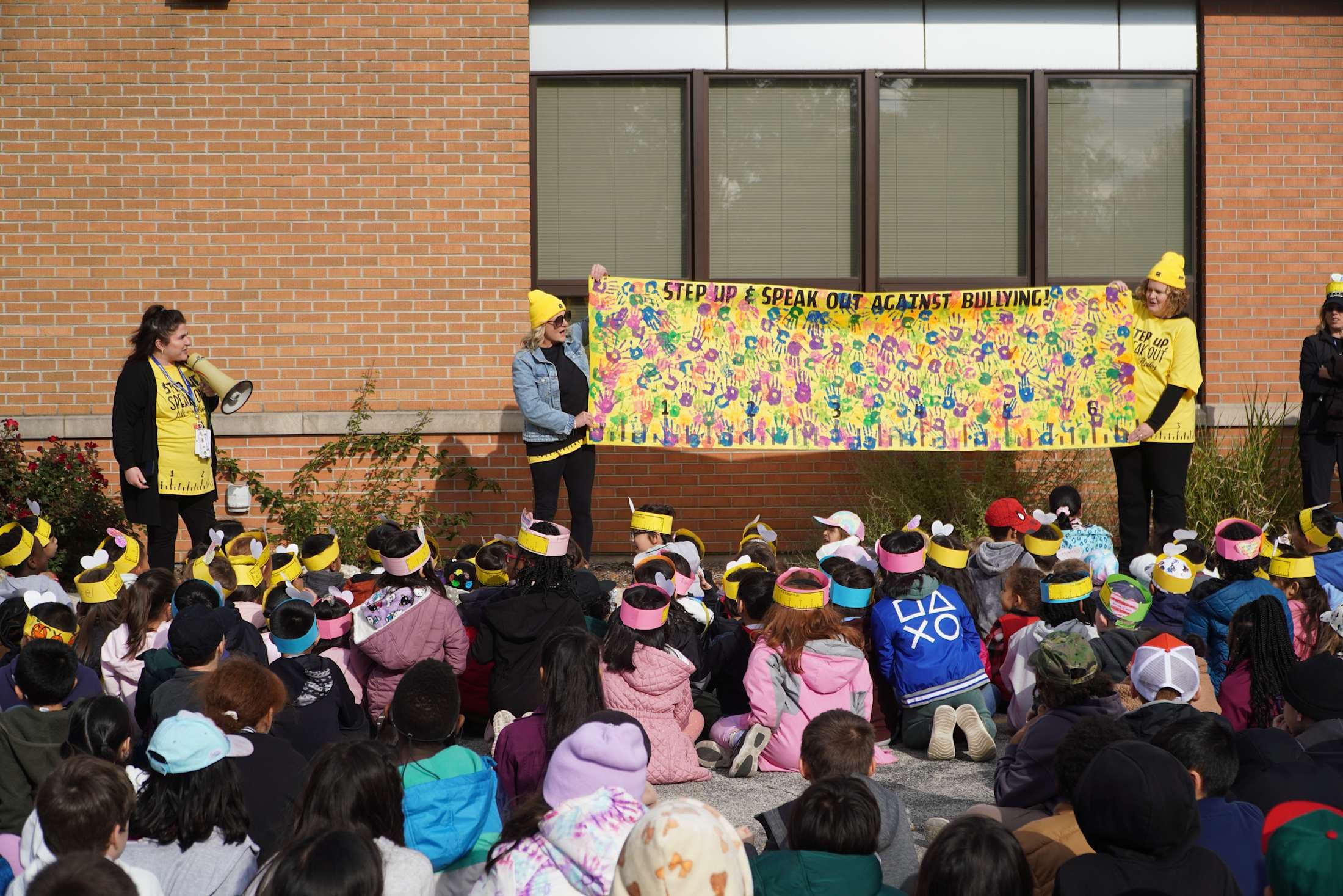 A group of children sit on the ground in the foreground while two adults hold a colorful anti-bullying banner in front of a brick building.