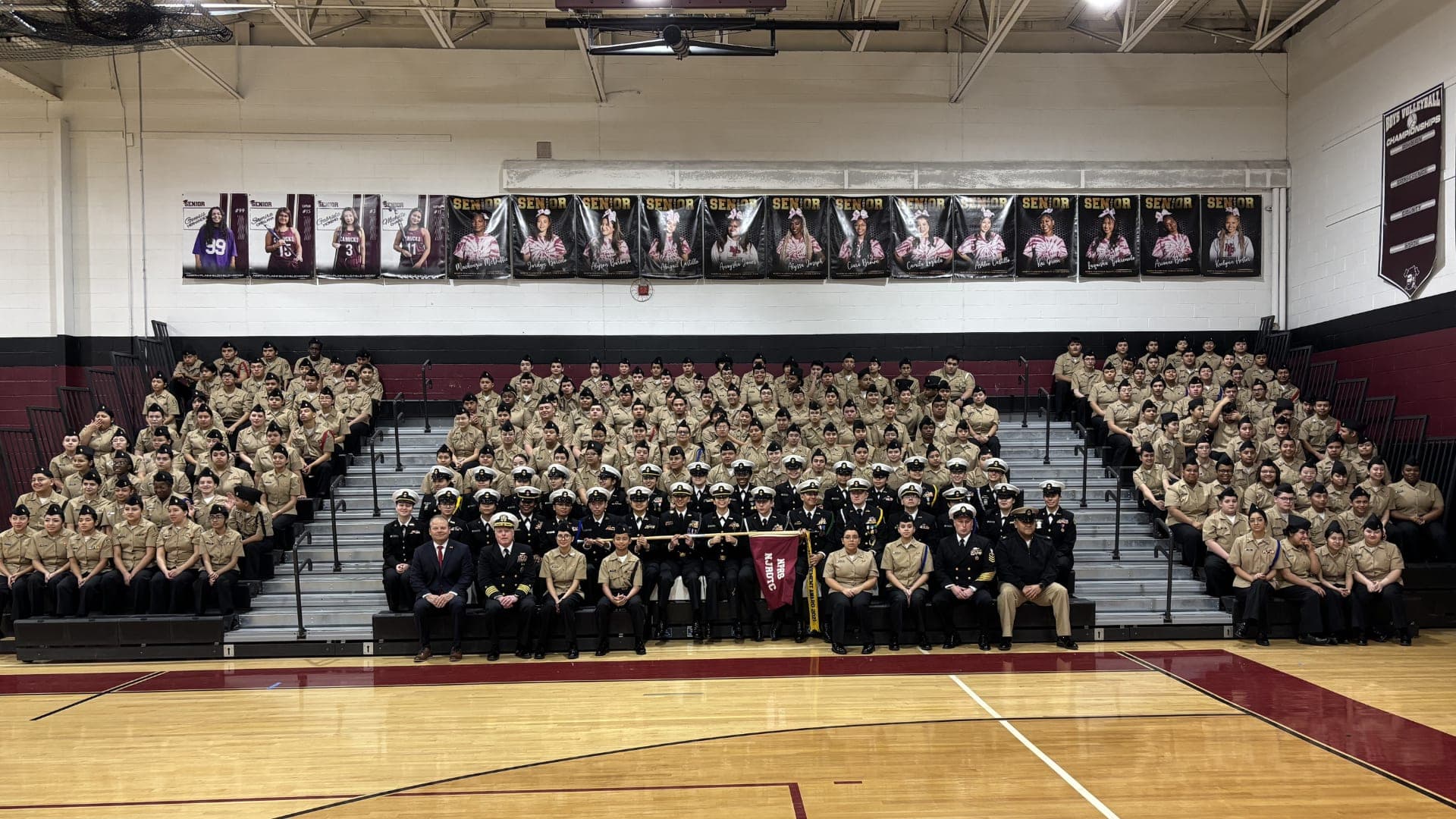A large group of people, likely a school or organization, gathered in a gymnasium setting, with banners and trophies displayed on the walls.
