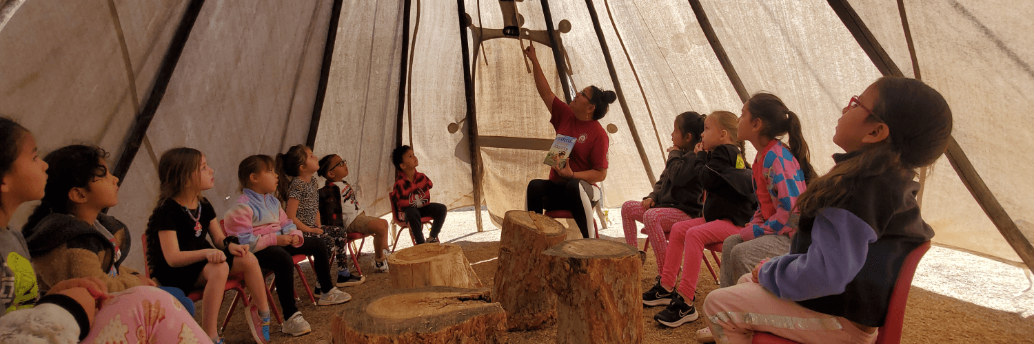 A group of people sit inside a teepee, possibly listening to a presentation.
