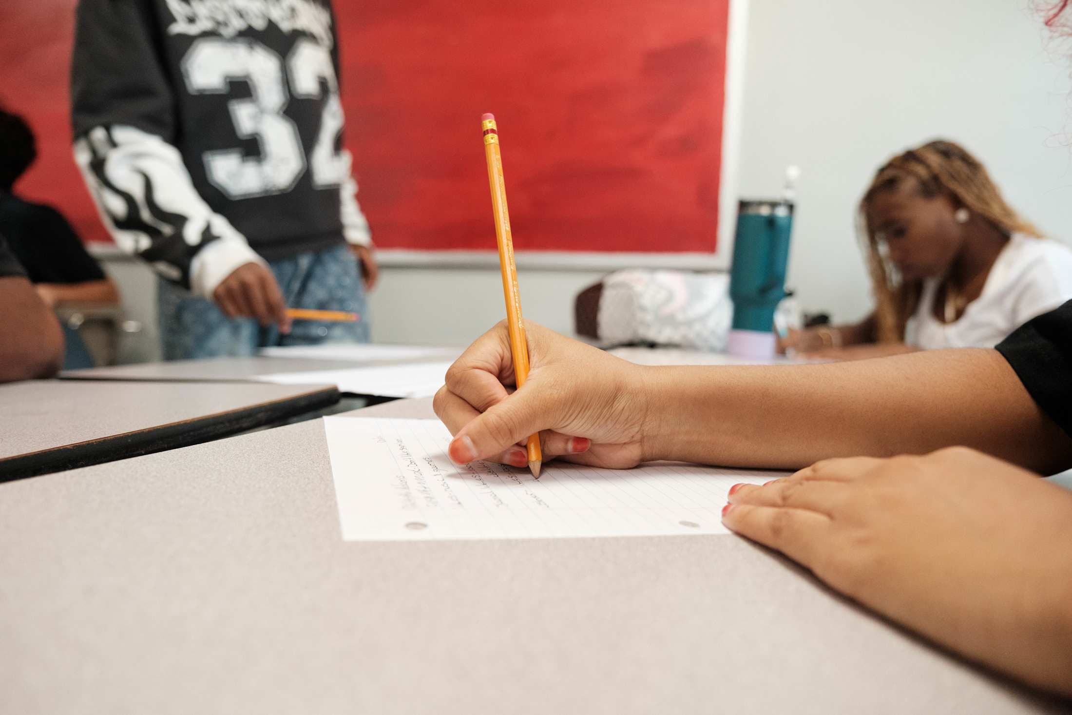 A person's hand writes with a pencil on a piece of paper in the foreground, while other students and a red background wall are visible behind them.
