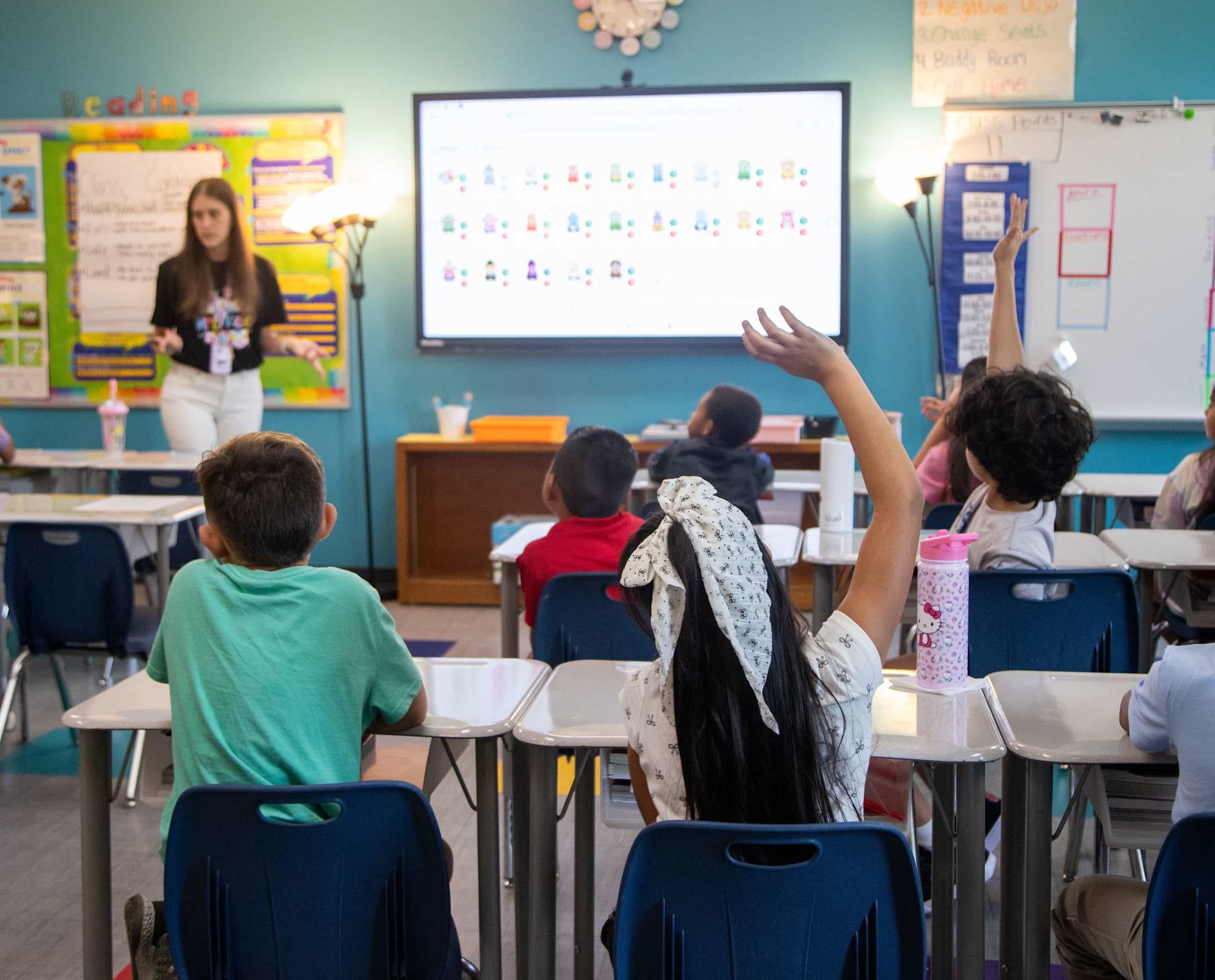 A teacher stands at the front of a classroom, gesturing towards a screen displaying musical notes, while students in the foreground raise their hands to answer questions.